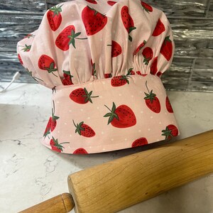 Puede incluir: Un gorro de chef rosa con estampado de fresas y lunares blancos. El gorro est&aacute; sobre una encimera blanca junto a un rodillo de madera.