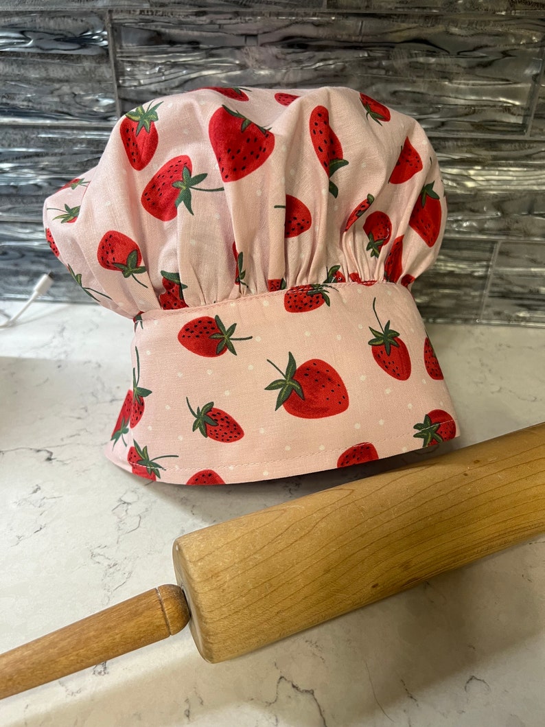 Puede incluir: Un gorro de chef rosa con estampado de fresas y lunares blancos. El gorro est&aacute; sobre una encimera blanca junto a un rodillo de madera.