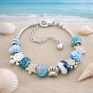 May include: Silver charm bracelet with various blue and silver beads. The beads include a turtle, starfish, and a bead with a pink rose design. The bracelet is on a sandy surface with seashells and the ocean in the background.