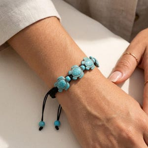 May include: A turquoise turtle charm bracelet with a black adjustable cord. The bracelet features several turquoise turtle charms and turquoise beads. The bracelet is on a wrist, with a neutral background.