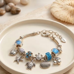 May include: A silver-tone charm bracelet featuring various ocean-themed pendants including two sea turtles, a starfish, a seashell, and a strawberry. The bracelet includes blue glass beads, blue crystal-encrusted charms, and a tag labeled Oceanic.
