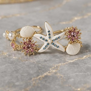 May include: Gold-toned rings with seashell, starfish, and crystal embellishments. The starfish is white with blue and pearl accents. The rings also feature pink and pearl-colored crystals.