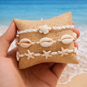 May include: Three beach-themed bracelets with white cord and gold accents. One bracelet features a turtle charm, another has seashells, and the third has starfish. The bracelets are displayed on a small, tan pillow with a blurred ocean background.
