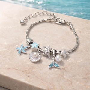 May include: Silver charm bracelet featuring ocean-themed charms. Charms include a starfish, a seashell with a pearl, a crystal-studded charm, a mermaid tail, and a star. The bracelet has a lobster clasp and a heart-shaped charm.
