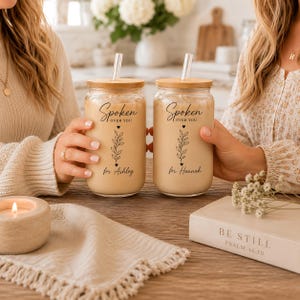 May include: Two glass tumblers with bamboo lids and clear straws, filled with a creamy beverage. Each tumbler is personalized with the text "Spoken Over You" and a name, "Ashley" and "Hannah". The tumblers are on a wooden table.