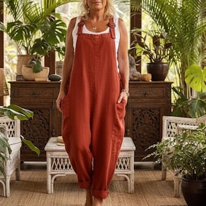 May include: A rust-colored jumpsuit with large front pockets and adjustable straps. The jumpsuit is worn over a white tank top. The person is standing barefoot in a sunlit room with plants and wicker furniture.