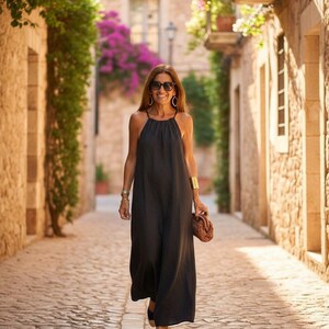 K&ouml;nnte beinhalten: Eine Frau in einem schwarzen Maxikleid mit Halfter, Sonnenbrille und Goldschmuck geht eine Kopfsteinpflasterstra&szlig;e entlang. Sie tr&auml;gt eine braune Clutch und Espadrilles. Die Szene spielt in einer europ&auml;ischen Stadt mit Steingeb&auml;uden und bl&uuml;henden Pflanzen.