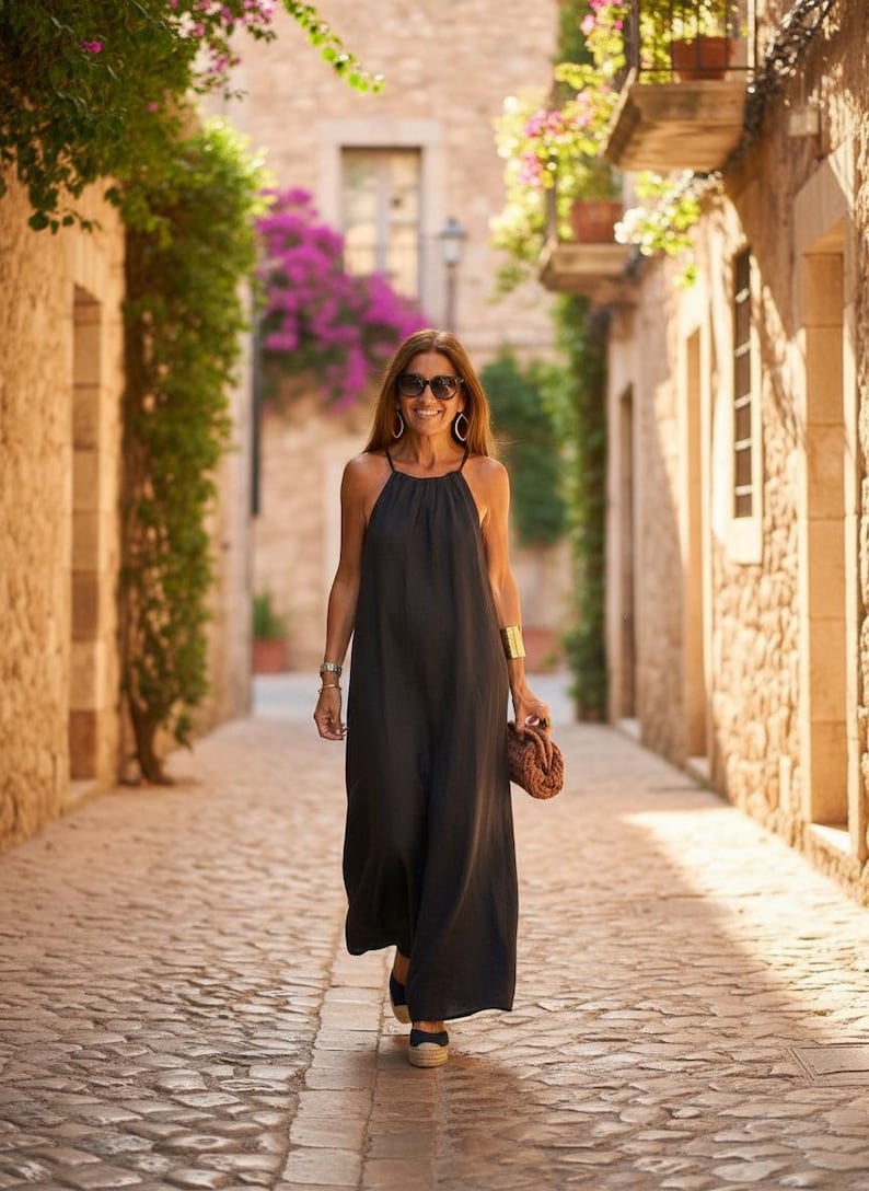 K&ouml;nnte beinhalten: Eine Frau in einem schwarzen Maxikleid mit Halfter, Sonnenbrille und Goldschmuck geht eine Kopfsteinpflasterstra&szlig;e entlang. Sie tr&auml;gt eine braune Clutch und Espadrilles. Die Szene spielt in einer europ&auml;ischen Stadt mit Steingeb&auml;uden und bl&uuml;henden Pflanzen.