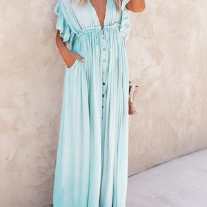 May include: A light blue maxi dress with a V-neckline and button-down front. The dress features short, ruffled sleeves, a drawstring waist, and pockets. The floor-length dress is made of a soft, flowing fabric.