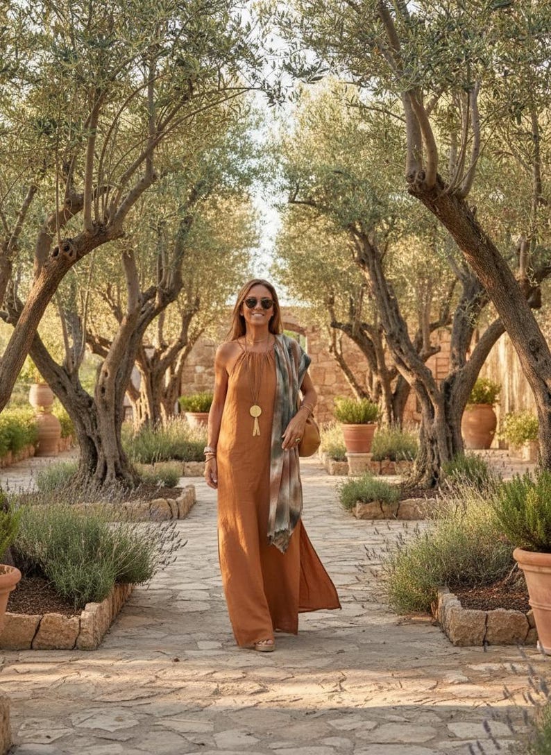 K&ouml;nnte beinhalten: Eine Frau in einem rostfarbenen Maxikleid mit Halfter-Ausschnitt, Sonnenbrille und einem gemusterten Schal geht einen Steinweg entlang, der von Olivenb&auml;umen und Topfpflanzen ges&auml;umt ist. Sie tr&auml;gt eine runde Tasche und eine goldene Anh&auml;ngerkette.