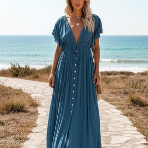 May include: A long, flowing teal dress with a deep V-neckline and short, flutter sleeves. The dress features a button-down front and a drawstring waist. The model is standing on a stone path near the ocean.