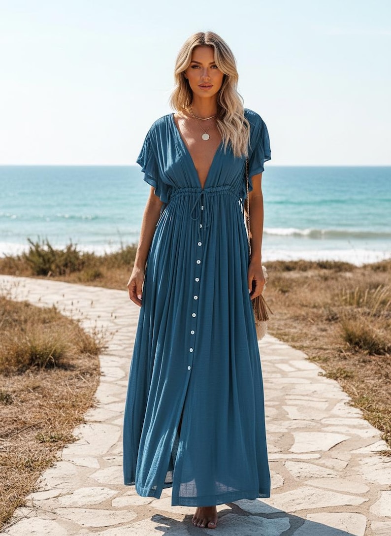 May include: A long, flowing teal dress with a deep V-neckline and short, flutter sleeves. The dress features a button-down front and a drawstring waist. The model is standing on a stone path near the ocean.