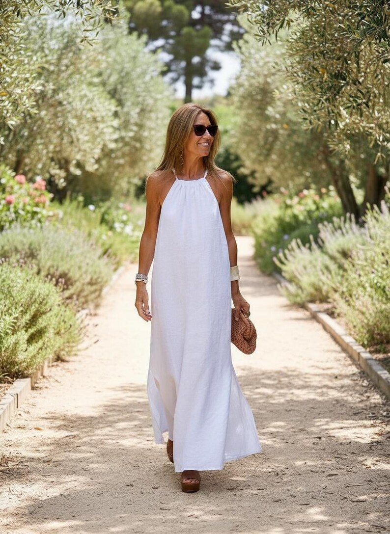 K&ouml;nnte beinhalten: Ein wei&szlig;es Maxikleid mit Neckholder-Ausschnitt. Das Kleid wird von einer Person getragen, die auf einem Weg geht. Die Person tr&auml;gt eine Sonnenbrille, ein Armband und eine gewebte Clutch. Der Hintergrund zeigt einen Garten mit B&auml;umen und Gr&uuml;n.