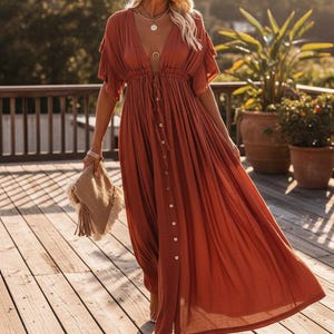 May include: Rust-colored maxi dress with a V-neckline, short flutter sleeves, and a button-down front. The dress features a drawstring waist and is worn with a woven clutch. The model is standing on a wooden deck.