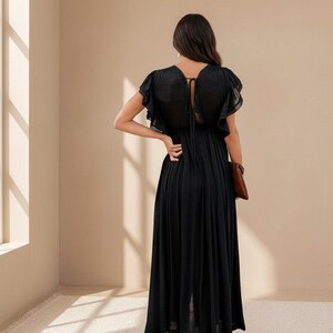 May include: A black maxi dress with short, ruffled sleeves and a tie detail at the back. The dress has a flowing skirt and is paired with a brown clutch. The model is standing near a window.