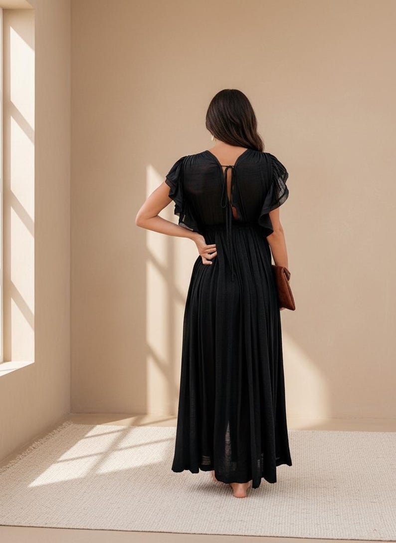 May include: A black maxi dress with short, ruffled sleeves and a tie detail at the back. The dress has a flowing skirt and is paired with a brown clutch. The model is standing near a window.