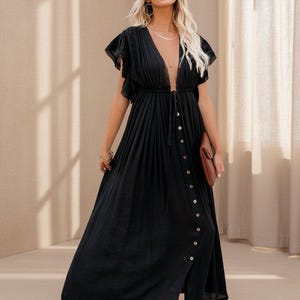 May include: A black maxi dress with a deep V-neckline and button-down front. The dress features short, ruffled sleeves and a flowing skirt. A brown clutch is held in the hand. The model is wearing gold jewelry.