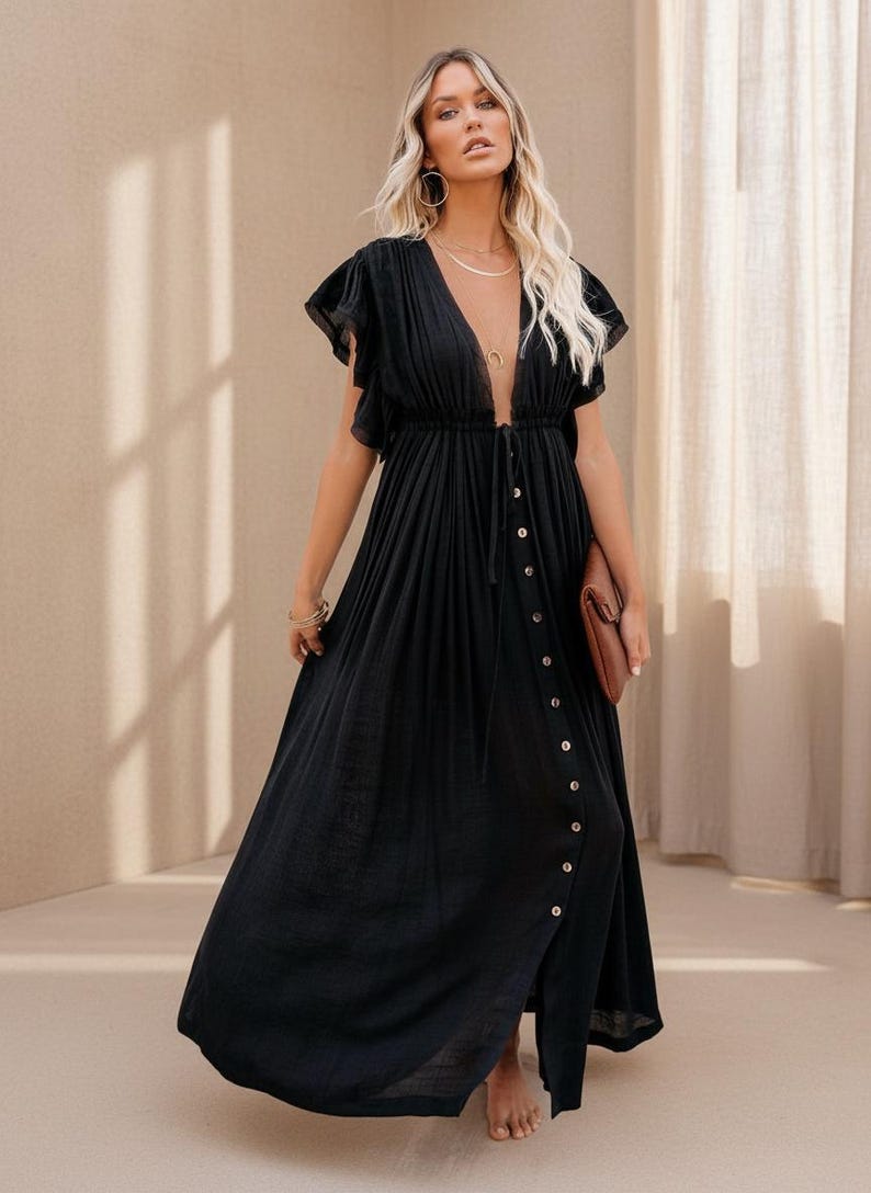May include: A black maxi dress with a deep V-neckline and button-down front. The dress features short, ruffled sleeves and a flowing skirt. A brown clutch is held in the hand. The model is wearing gold jewelry.
