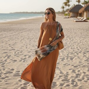 K&ouml;nnte beinhalten: Eine Person tr&auml;gt ein langes, flie&szlig;endes, rostfarbenes Kleid und eine Sonnenbrille an einem Sandstrand. Sie tr&auml;gt eine braune Handtasche und einen gemusterten Schal. Eine goldene Anh&auml;ngerkette und Sandalen vervollst&auml;ndigen den Look. Palmen und Strandh&uuml;tten sind im Hintergrund.