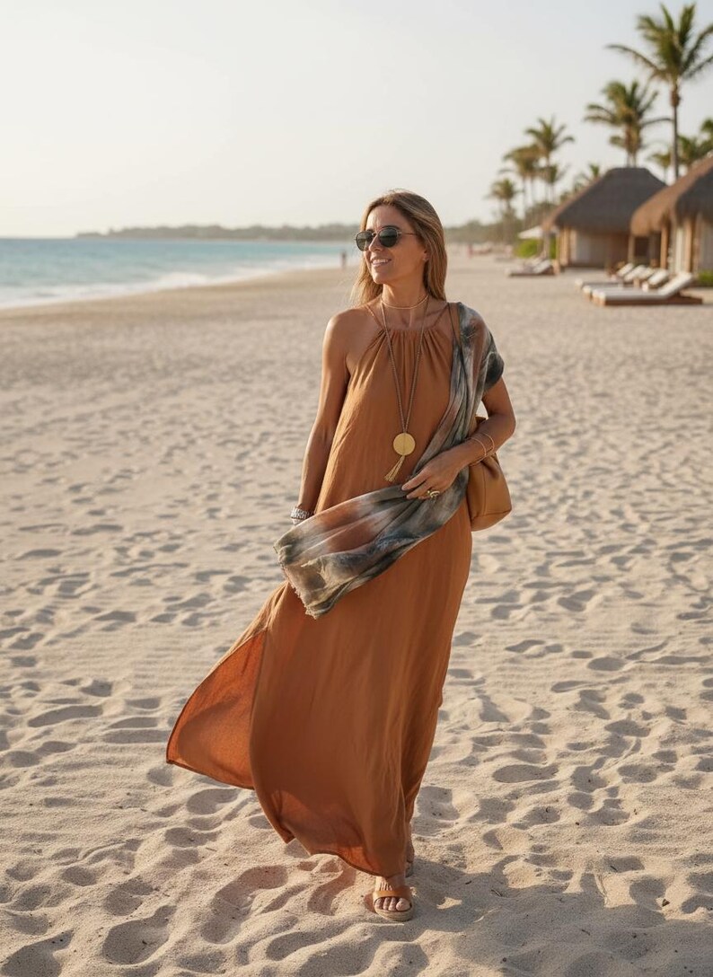 K&ouml;nnte beinhalten: Eine Person tr&auml;gt ein langes, flie&szlig;endes, rostfarbenes Kleid und eine Sonnenbrille an einem Sandstrand. Sie tr&auml;gt eine braune Handtasche und einen gemusterten Schal. Eine goldene Anh&auml;ngerkette und Sandalen vervollst&auml;ndigen den Look. Palmen und Strandh&uuml;tten sind im Hintergrund.