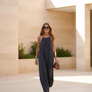 K&ouml;nnte beinhalten: Ein dunkelgraues Maxikleid mit Halterneck. Das Kleid wird mit goldenen Armb&auml;ndern, Creolen, Sonnenbrille und einer braunen Clutch kombiniert. Die Person tr&auml;gt blaue Espadrilles. Der Hintergrund ist ein hellbeige Geb&auml;ude.