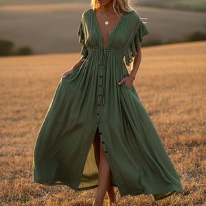 May include: A flowing, olive green maxi dress with a deep V-neckline and button-down front. The dress features short, ruffled sleeves, a drawstring waist, and pockets. The model is standing in a field of dry grass.