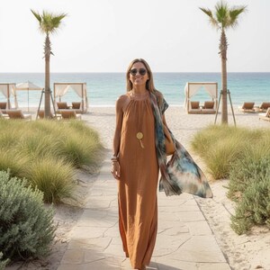 K&ouml;nnte beinhalten: Eine Person in einem langen, rostfarbenen Kleid mit Halfter-Ausschnitt und Sonnenbrille geht auf einem Steinweg zum Strand. Sie tr&auml;gt einen gemusterten Schal und eine goldene Anh&auml;ngerkette. Palmen und Strandh&uuml;tten sind im Hintergrund zu sehen.