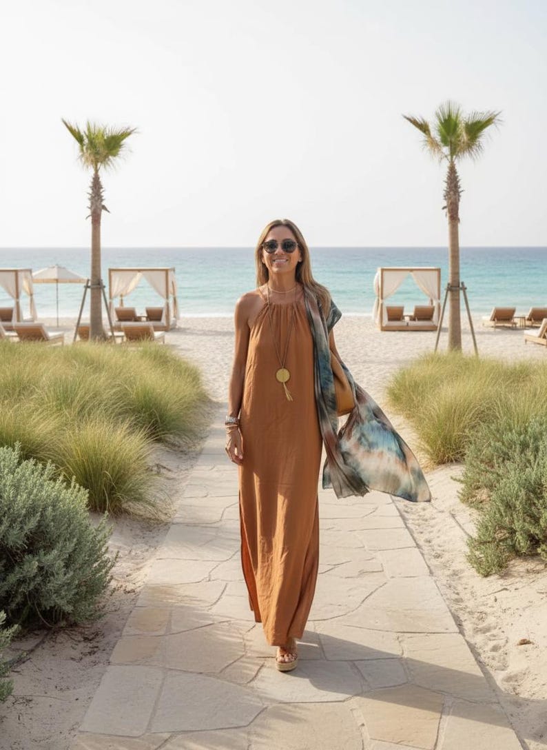 K&ouml;nnte beinhalten: Eine Person in einem langen, rostfarbenen Kleid mit Halfter-Ausschnitt und Sonnenbrille geht auf einem Steinweg zum Strand. Sie tr&auml;gt einen gemusterten Schal und eine goldene Anh&auml;ngerkette. Palmen und Strandh&uuml;tten sind im Hintergrund zu sehen.