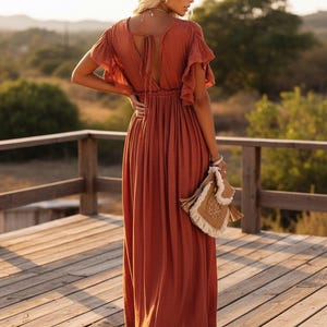 May include: Rust-colored maxi dress with flutter sleeves and a V-neckline. The dress features a tie detail at the back and a gathered waist. A woven handbag with fringe is held in the hand. The dress is worn on a wooden deck.
