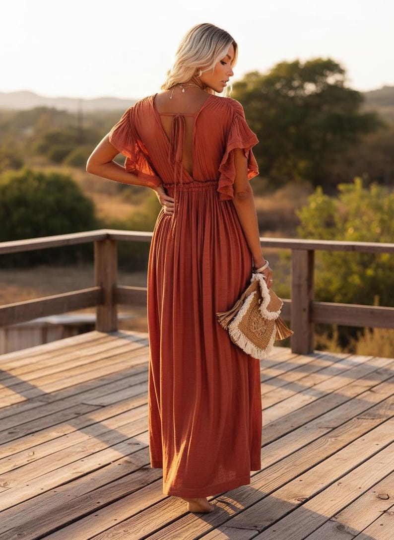 May include: Rust-colored maxi dress with flutter sleeves and a V-neckline. The dress features a tie detail at the back and a gathered waist. A woven handbag with fringe is held in the hand. The dress is worn on a wooden deck.
