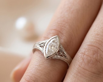 May include: A silver-toned ring featuring a central marquise-cut clear gemstone set in a bezel. The band displays intricate openwork metal detailing with a delicate milgrain edge, creating a refined and structured appearance on the finger.