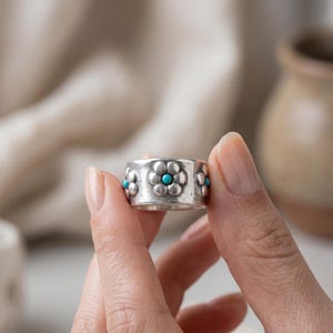 May include: A wide silver band ring featuring a repeating embossed flower pattern. Each flower has a small, round, bright turquoise stone set in the center. The metal has a slightly weathered, matte finish with visible texture across the surface.