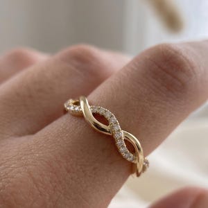 May include: A gold ring featuring an intertwined infinity design. One strand is polished gold, while the other is encrusted with small, clear, round-cut stones that catch the light. The band has a continuous, flowing, twisted shape.