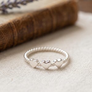 May include: A silver ring featuring a twisted band design with three small, smooth, polished heart-shaped charms attached to the front. The metal has a bright, reflective finish, and the band consists of a continuous rope-like texture.