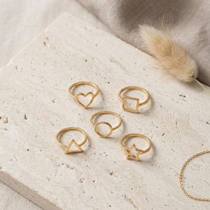 Peut inclure: Un ensemble de cinq bagues dorées, chacune présentant une forme géométrique ou symbolique distincte. La collection comprend un cœur, un carré, un cercle, un triangle et une étoile, tous conçus avec des anneaux fins et une finition métallique.