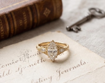 May include: A gold ring featuring a central marquise-cut gemstone flanked by two smaller marquise-cut stones on each side. The band displays intricate metalwork detailing along the shoulders, providing a textured appearance to the gold setting.