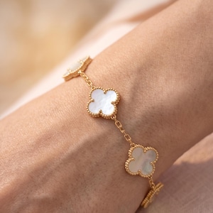 May include: A gold chain bracelet featuring multiple four-leaf clover motifs. Each clover charm is outlined with a beaded gold edge and contains a smooth, iridescent white mother-of-pearl inlay. The bracelet is secured with a delicate gold link chain.