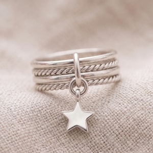 May include: A silver ring featuring a stack of four bands, alternating between two smooth bands and two twisted rope-textured bands. A small, polished silver star charm hangs from a vertical loop attached to the center of the ring stack.