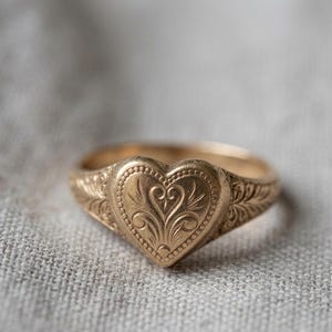 May include: A gold ring featuring a heart-shaped centrepiece with an engraved floral pattern. The band and the heart are decorated with intricate, fine line details and a beaded border, creating a textured surface across the entire metal piece.