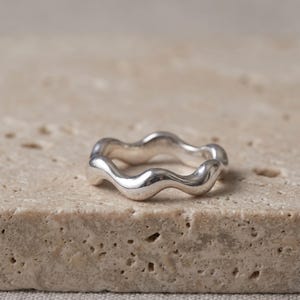Puede incluir: Un anillo de plata pulida que presenta un diseño continuo, fluido y ondulado. La banda tiene una superficie metálica lisa y reflectante con un patrón ondulante repetitivo que crea una serie de curvas suaves alrededor de toda la pieza.