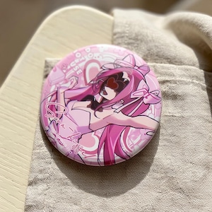 Op de afbeelding: Een ronde button met een levendige roze anime-illustratie van een personage met lang roze haar, grote strikken en een hartvormige bril. Het ontwerp bevat witte tekst, hartmotieven en een glanzende afwerking op het oppervlak van de button.