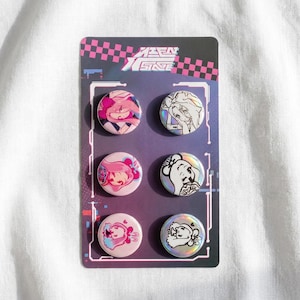 Op de afbeelding: Een set van zes ronde buttons op een rechthoekige kaart met een paars en zwart geblokt patroon. De buttons tonen anime-stijl karakterillustraties in roze, blauw en wit, waarbij sommige een holografische afwerking hebben.