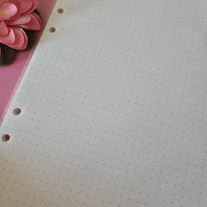 K&ouml;nnte beinhalten: Ein Blatt cremefarbenes Punktrasterpapier mit drei L&ouml;chern am linken Rand. Das Papier ist mit kleinen, gleichm&auml;&szlig;ig verteilten grauen Punkten bedeckt. Eine rosa Blume ist in der oberen linken Ecke zu sehen.
