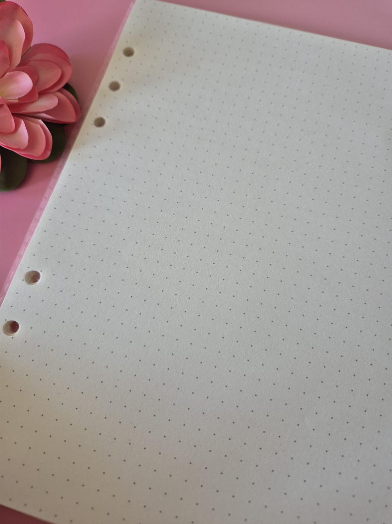 K&ouml;nnte beinhalten: Ein Blatt cremefarbenes Punktrasterpapier mit drei L&ouml;chern am linken Rand. Das Papier ist mit kleinen, gleichm&auml;&szlig;ig verteilten grauen Punkten bedeckt. Eine rosa Blume ist in der oberen linken Ecke zu sehen.