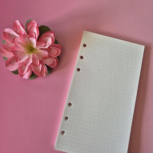 May include: A pink paper flower with green leaves sits next to a white dot grid paper with six holes on a pink background. The paper is blank and ready for use. The flower has multiple layers of pink petals.