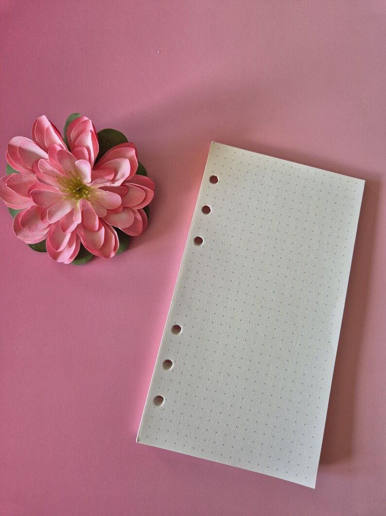 May include: A pink paper flower with green leaves sits next to a white dot grid paper with six holes on a pink background. The paper is blank and ready for use. The flower has multiple layers of pink petals.