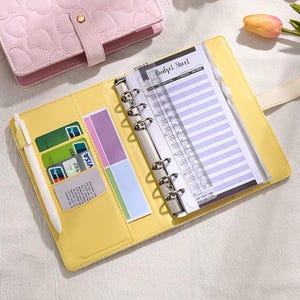 May include: Open yellow planner with a silver binder, holding a budget sheet and a pen. The planner has card slots with credit cards and colorful sticky notes. A pink heart-patterned planner is visible in the background.