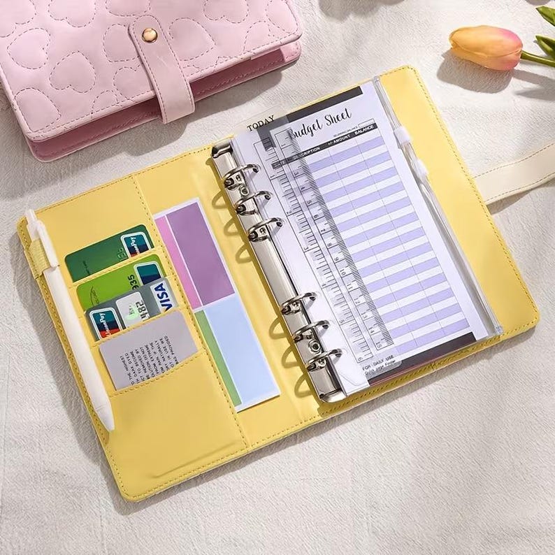 May include: Open yellow planner with a silver binder, holding a budget sheet and a pen. The planner has card slots with credit cards and colorful sticky notes. A pink heart-patterned planner is visible in the background.