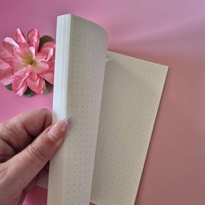 May include: A dot grid notebook with pages being turned, revealing the interior pages. The notebook is held by a hand with a pink flower in the background. The pages have a dot grid pattern.