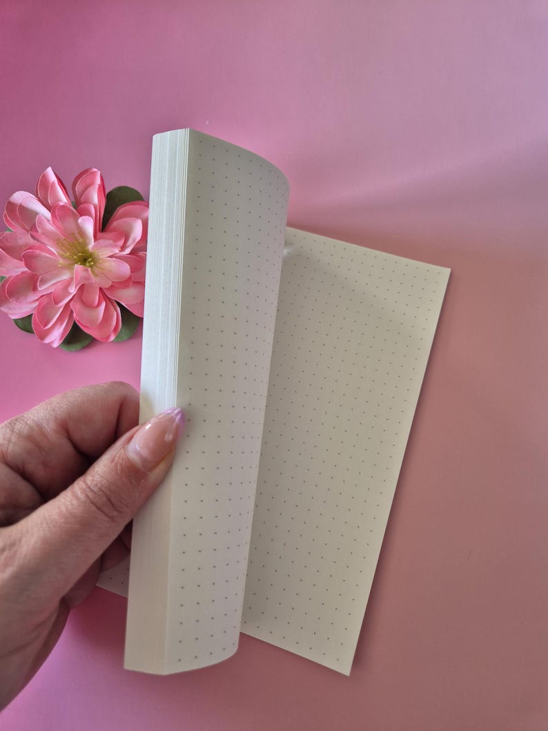 May include: A dot grid notebook with pages being turned, revealing the interior pages. The notebook is held by a hand with a pink flower in the background. The pages have a dot grid pattern.